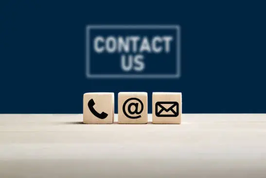 Wooden blocks showing phone, at-sign, and mail icons on a desk with a blurred Contact Us sign, representing ways to reach a digital marketing and SEO consultant