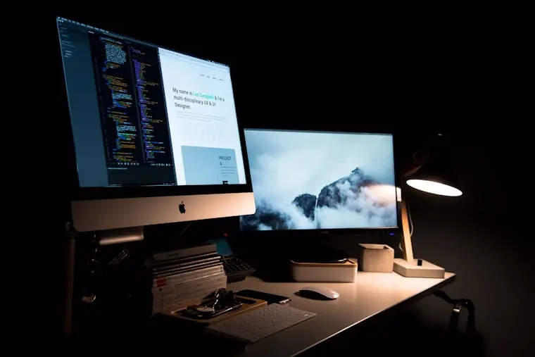 A professional dual-monitor desk setup displaying website source code and a live homepage design, illustrating custom WordPress web development and design services